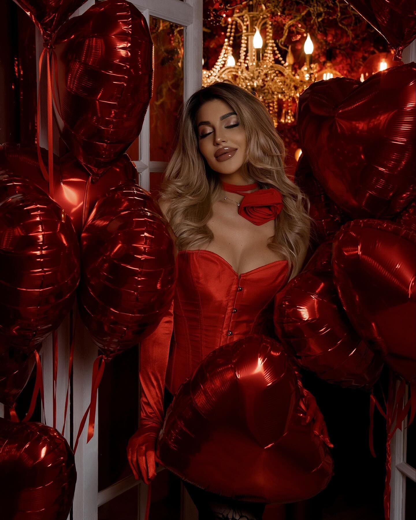 A woman with eyes closed stands among red heart balloons wearing a red satin corset top and choker.