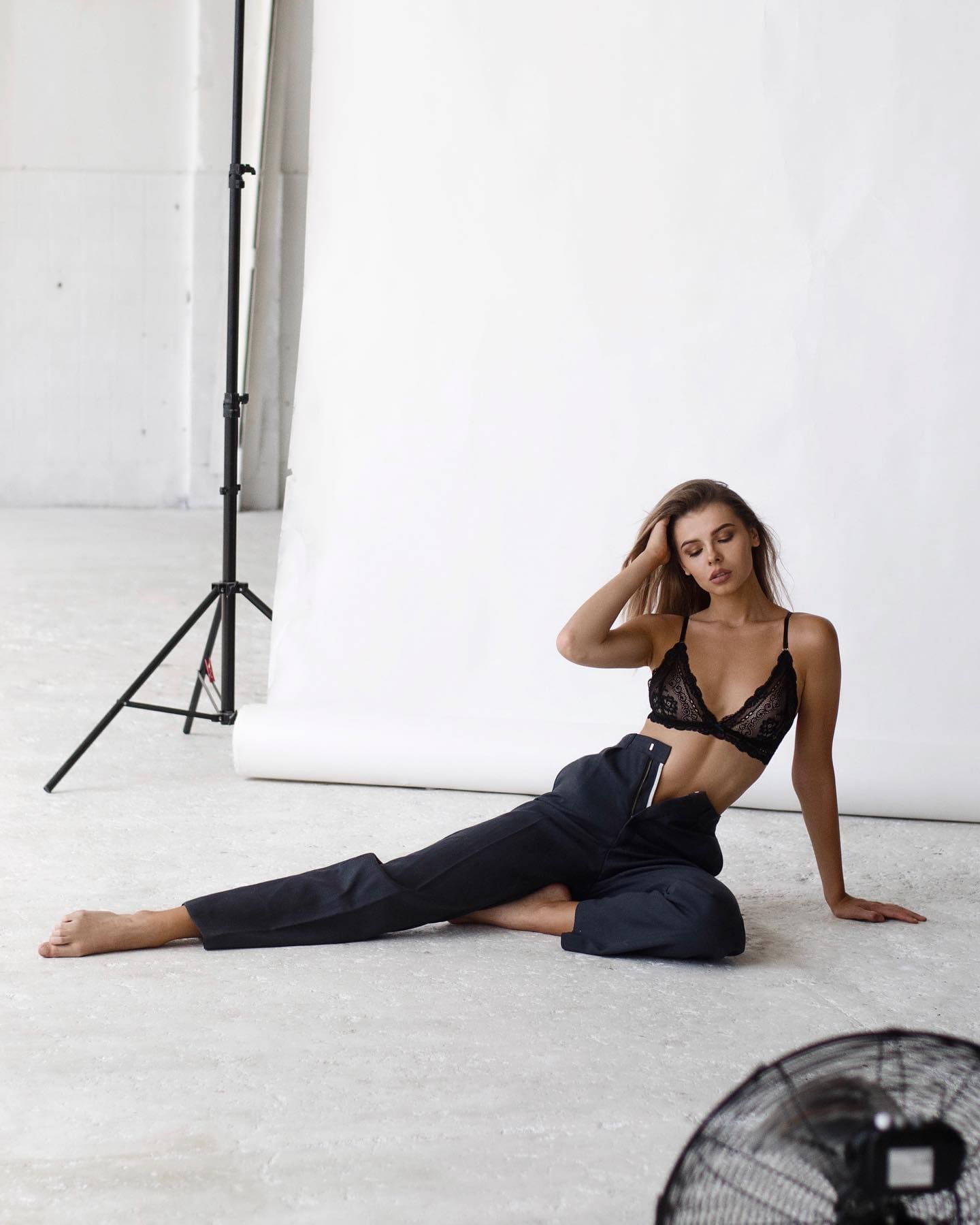 A woman with brown hair sits on a concrete floor in a black lace bra and trousers, hand in hair, with a lighting stand behind.