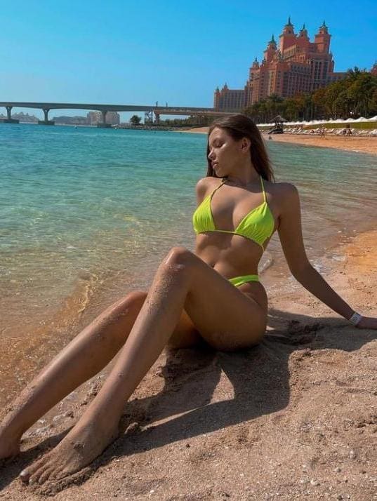 A woman in a yellow bikini sits on a beach with legs extended, an ornate building and bridge visible in the background.