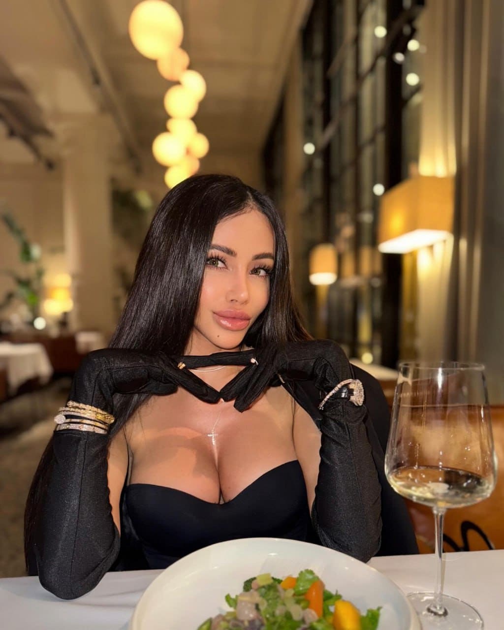 A woman sits at a restaurant table in a black strapless top and jacket, hands near her shoulders, with a salad plate and wine glass in front.