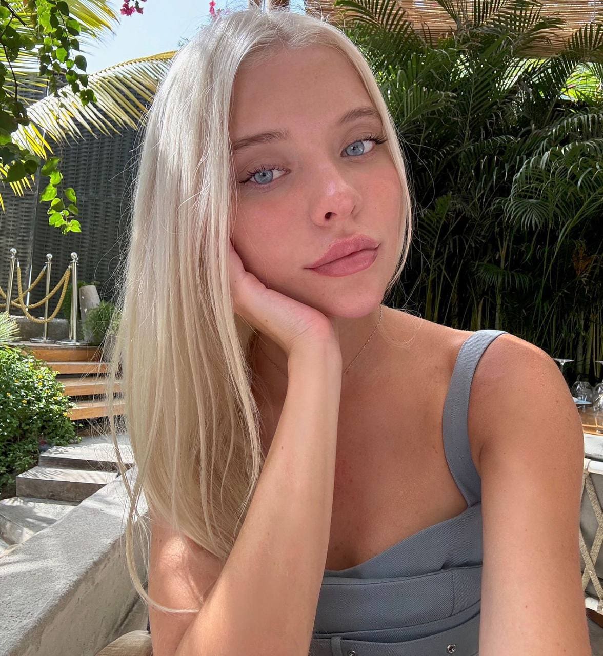 Seated outdoors, a platinum blonde rests her chin on her hand, wearing a grey low-neck top before green palms.