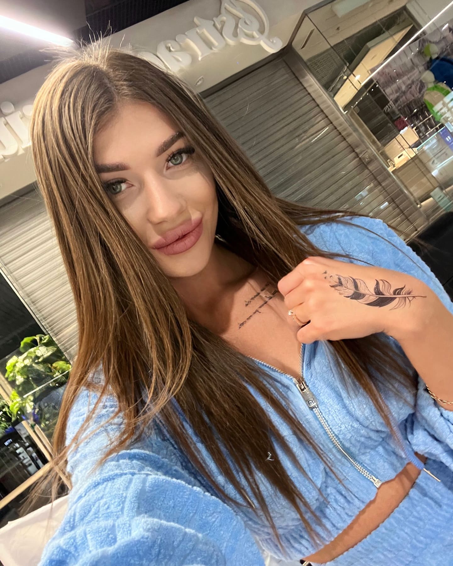 A selfie of a woman in a light blue zip-up top, showing hand and chest tattoos, with a storefront visible in the background.
