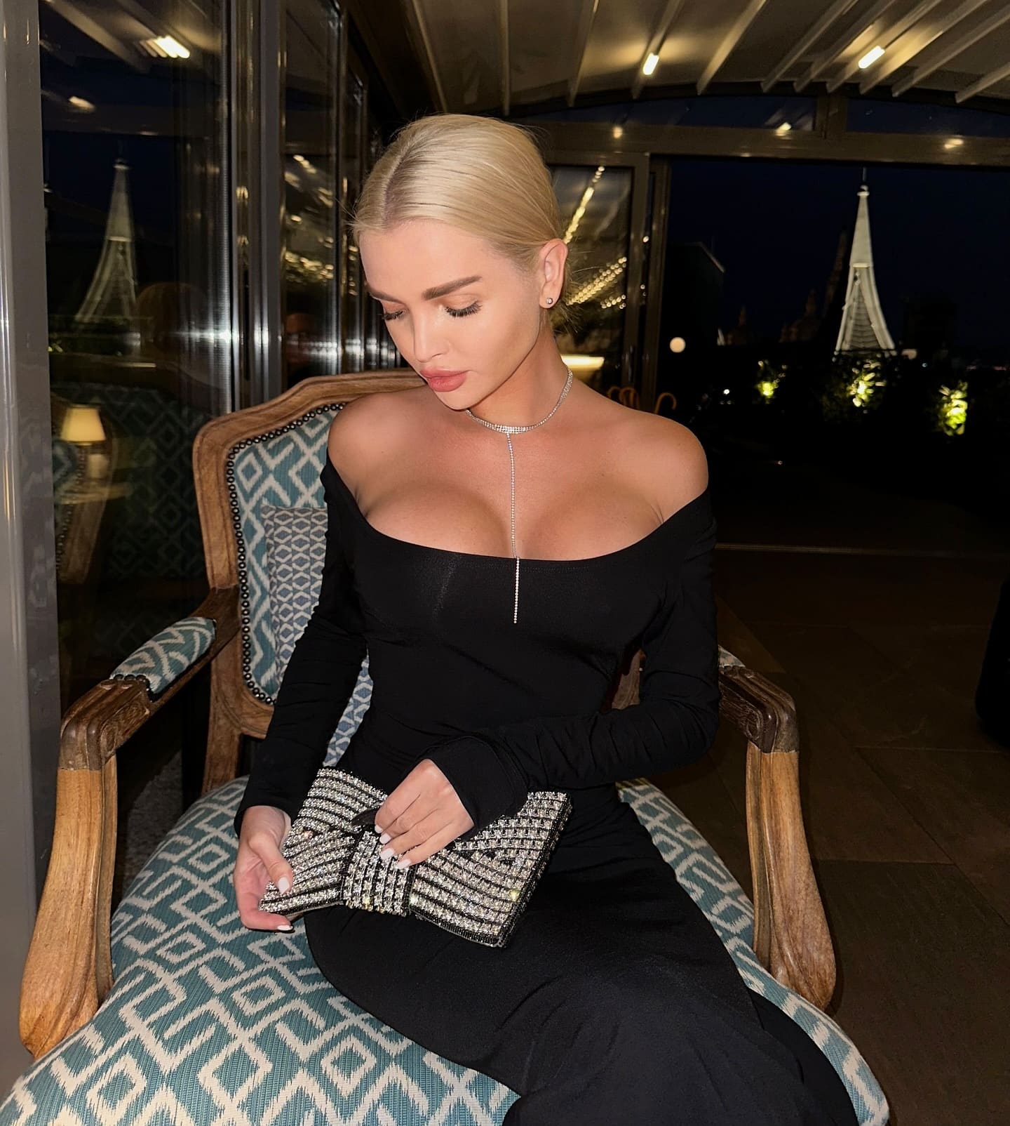 A blonde woman sits in a patterned armchair wearing a black off-the-shoulder dress, holding a beaded clutch near large windows.